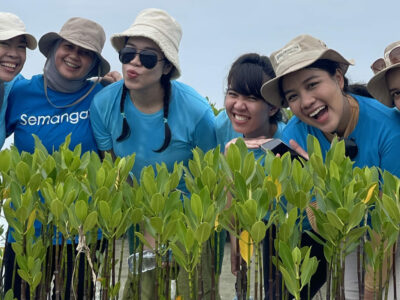 6 wanita berbaju biru Bumi Journey dengan bucket hat sedang gathering kantor berdiri di belakang bibit mangrove di pesisir pantai