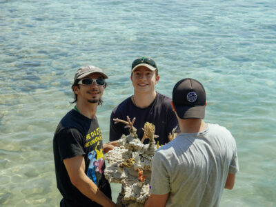 3 pria peserta acara penanaman terumbu karang bersama mengangkat benih terumbu karang yang akan ditanam dalam upaya mencapai SDGs