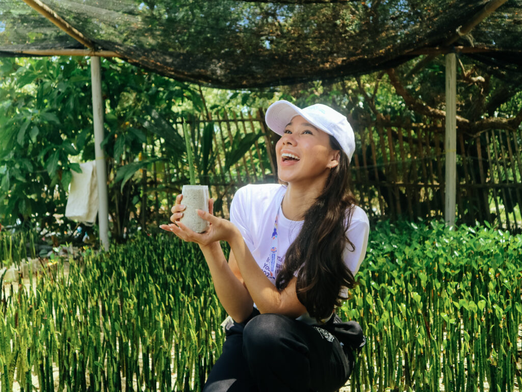 wanita bertopi putih dan berbaju putih tersenyum lembar melihat ke atas sambil memegang bibit mangrove di mangrove nursery
