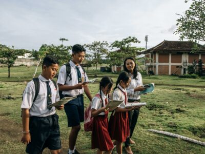 2 anak perempuan SD dan dua lelaki SMP berseragam belajar di ruang terbuka suasana pinggiran kota hijau bersama seorang guru wanita sambil memegang buku