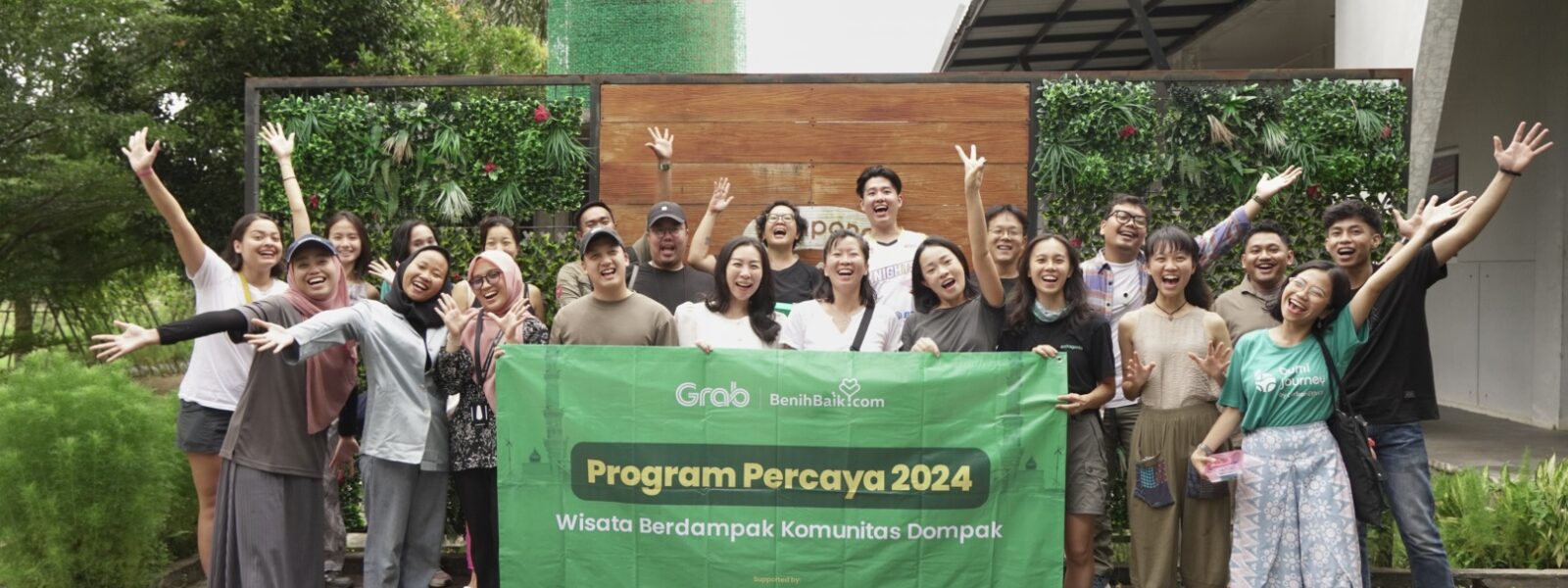 Kelompok peserta Program Percaya 2024 berpose ceria bersama sambil memegang spanduk bertuliskan "Wisata Berdampak Komunitas Dompak", di depan area hijau yang asri. Gambar ini menggambarkan contoh event organizer yang sukses menyelenggarakan acara wisata berdampak positif.