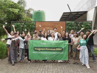 Kelompok peserta Program Percaya 2024 berpose ceria bersama sambil memegang spanduk bertuliskan "Wisata Berdampak Komunitas Dompak", di depan area hijau yang asri. Gambar ini menggambarkan contoh event organizer yang sukses menyelenggarakan acara wisata berdampak positif.