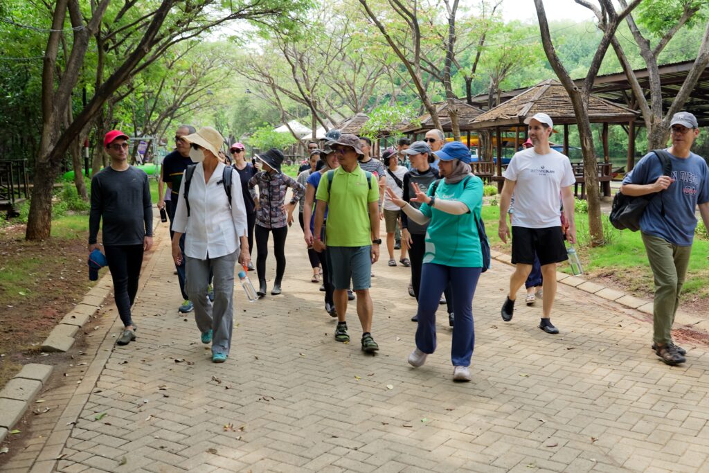 Sekelompok peserta berjalan santai di taman yang rindang, dipandu oleh seorang pemandu acara menggunakan kaos hijau. Kegiatan ini merupakan contoh event organizer yang mengadakan acara ramah lingkungan dan sehat di ruang terbuka.