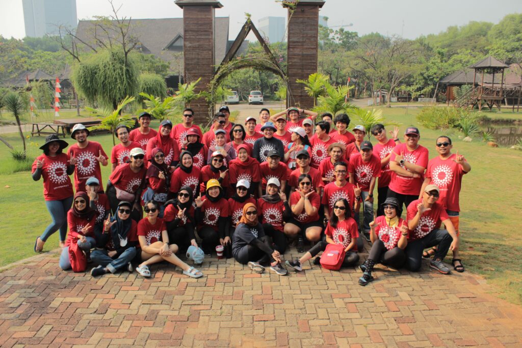 Foto kelompok besar peserta kegiatan outdoor mengenakan kaos merah seragam, berpose bersama di area taman dengan latar belakang hijau dan struktur kayu. Ini adalah contoh event organizer dalam mengelola acara team building untuk perusahaan.
