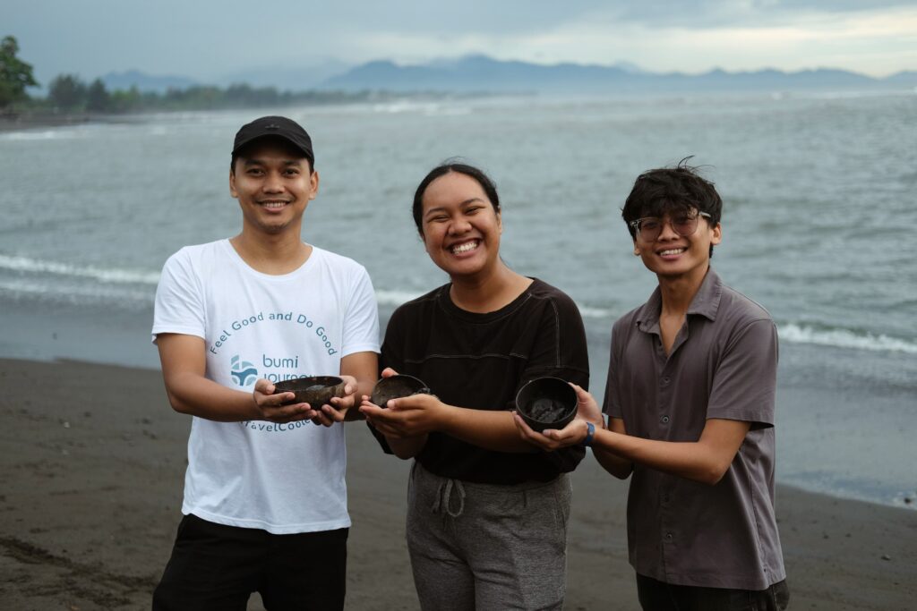 staf Bumi Journey berfoto dengan 2 wisatawan klien sambil memegang kerajinan lokal berupa mangkuk berwarna gelap di tepi pantai. Ekowisata adalah salah satu cara melestarikan kerajinan lokal ini.